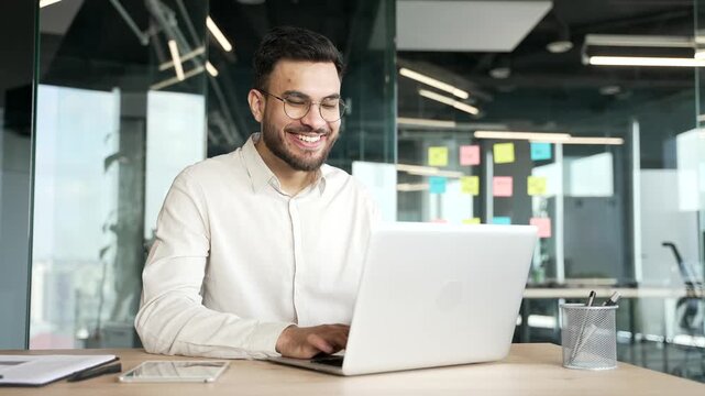 Satisfied happy businessman suit received great news on laptop while sitting at work desk at workplace in business office. Smiling glad handsome entrepreneur reads positive good message on computer