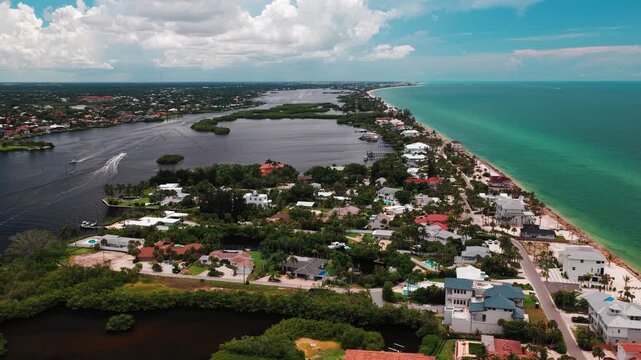 Residential peninsula sits between a dark tidal bay and bright turquoise gulf as a speedboat leaves a white wake, daytime drone flyover.