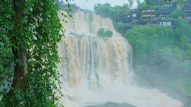Static wide shot of Furong waterfall cascading past traditional stilt houses with tree framing