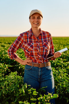 Smiling woman farmer standing in an agricultural field, holding a digital tablet and checking crop health