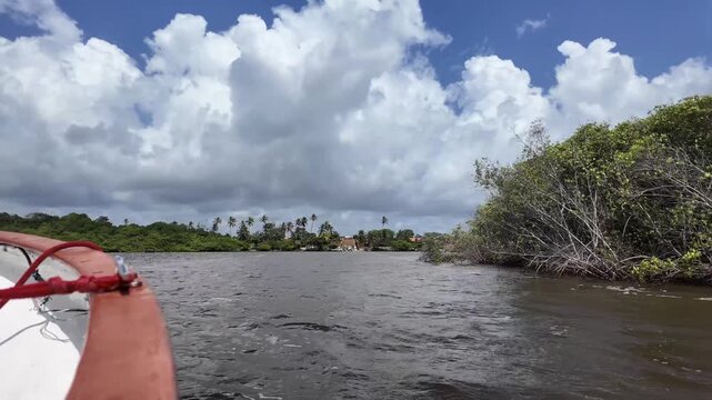 Scenic boat view across the Pregui&ccedil;as River toward Mandacaru village and its striped lighthouse beneath dramatic tropical clouds in Maranh&atilde;o, Brazil.