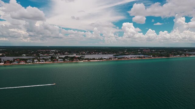 A white speedboat zips along turquoise gulf water in front of a long strip of houses and sandy shoreline under tall summer clouds.