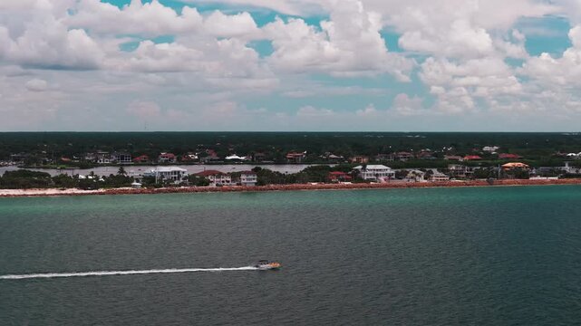 A small motorboat leaves a white wake across teal bay water in front of rock armored Osprey waterfront houses under puffy clouds.
