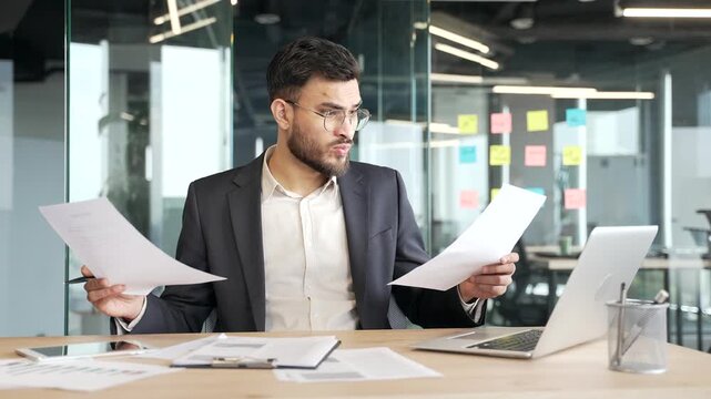 Confused businessman in a formal suit having difficulty with paperwork sitting at desk at workplace in modern business office. Puzzled entrepreneur looks at documents and cannot understand the problem
