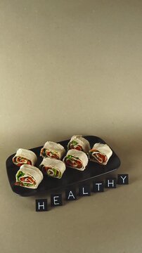 Delicious vegetable and ham lavash pinwheel rolls on a black plate with the word healthy spelled out, representing a nutritious snack, a balanced diet, and wellness on a plain background