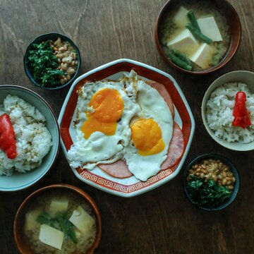 Top-down flat lay view of a traditional Japanese breakfast table spread featuring fried eggs on ham, bowls of white rice topped with mentaiko cod roe, natto with greens, and tofu miso soup.
