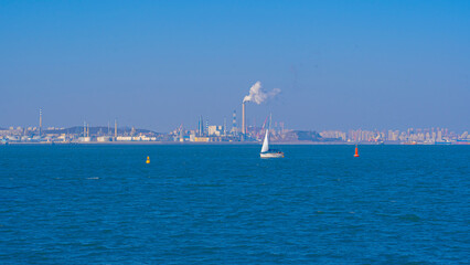 Horizontal view of a white sailboat on blue sea with industrial port and smoking chimney background in Dalian © 以诺 张