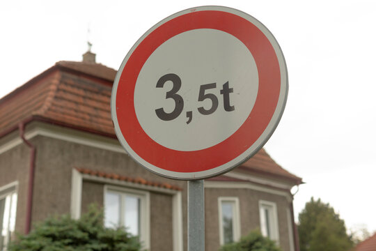 Weight limit sign in suburb closeup, circular red and white plate mounted on metal pole, brick house and trimmed hedge in background, overcast sky, municipal