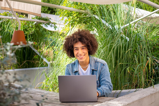 Smiling young ethnic guy 20 years old, dressed in casual clothes, works on a laptop outdoors, in a green area in nature. Green open space for coworking, freelancer, relaxing, coffee, food. Free place 
