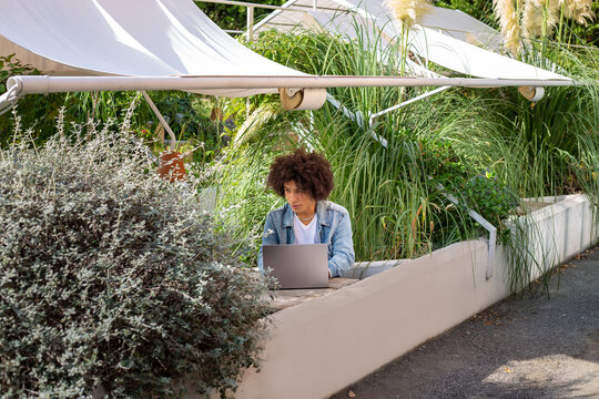 Smiling young ethnic guy 20 years old, dressed in casual clothes, works on a laptop outdoors, in a green area in nature. Green open space for coworking, freelancer, relaxing, coffee, food. Free place 