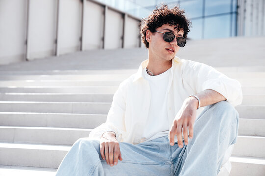 Young attractive man with curly hair hairstyle. Smiling  handsome male in casual stylish clothes posing in the street it sunny day. Cheerful and happy model outdoors in sunglasses, sits at stairs
