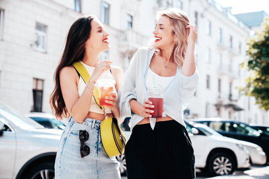 Two young beautiful smiling glam female in trendy summer clothes. Sexy women posing outdoors. Positive models holding and drinking fresh cocktail smoothie drink in plastic cup. Bright makeup, red lips