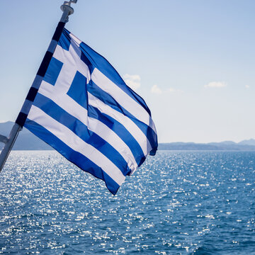 Greek flag billowing from ship above sparkling Aegean Sea water, blue sky, Greece