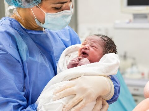 a crying newborn baby held by Medical worker in protective gown and face mask in a hospital delivery room during the first moments of postnatal care after childbirth.