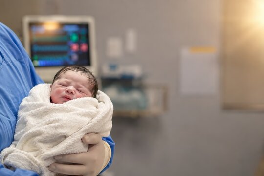 Newborn baby sleeps wrapped in a white hospital blanket while held by a healthcare worker in a maternity ward, with patient monitor in soft focus and generous copy space for medical messaging.