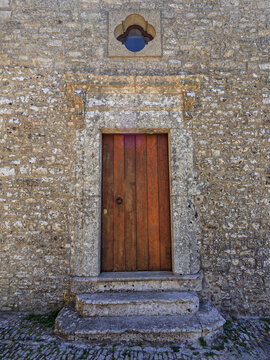 Vertical composition highlighting the weathered wood grain, lichen-covered stone surround, rustic fieldstone texture, and stone entry steps in Erice.
