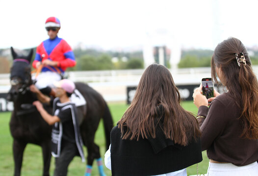 P&uacute;blico en hip&oacute;dromo, mujeres espectadoras con m&oacute;vil, caballo y jockey de carreras
