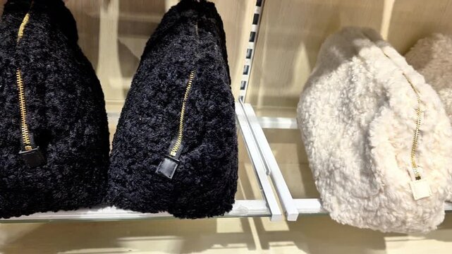 Soft textured handbags displayed on a retail shelf in a fashion store. Black and cream fluffy shoulder bags with gold-tone zippers arranged neatly for sale in a modern shopping environment.
