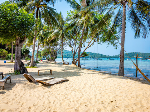 Alano Beach Karimunjawa showcasing a serene tropical landscape with golden sand, swaying palm trees, beach chairs, swings, and a hammock beside the clear blue ocean