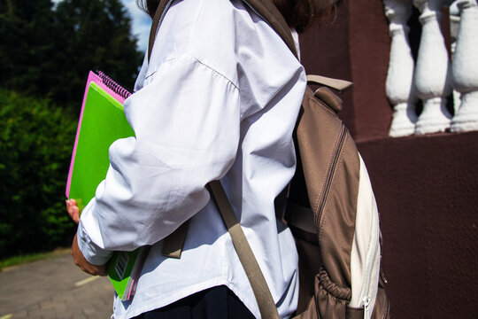 Schoolgirl stands in front of a school holding notebooks and supplies while wearing a backpack. It is back to school season