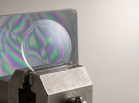 Curved sapphire watch crystal clamped in a metal vise under polarized light revealing colorful stress patterns