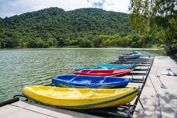 Kayaks Lined Up by the Lake: A vibrant row of colorful kayaks rests on a wooden dock, ready for adventure on a calm lake with a lush, tree-covered mountain in the background. © bubbers
