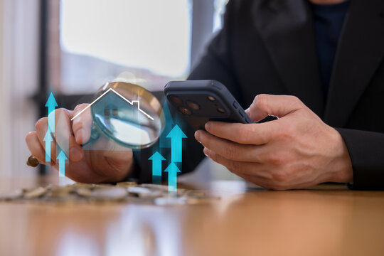 Businessperson using smartphone with magnifying glass over coins, house icon rising arrows, representing real estate investment, property market growth finance mortgage home valuation.