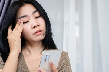 Asian woman suffering from eye strain and dry eyes touching her face while looking at phone. Ideal...