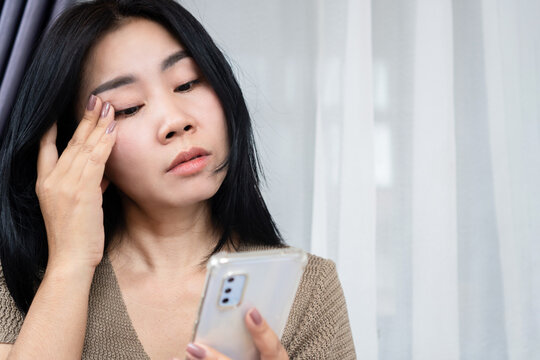 Asian woman suffering from eye strain and dry eyes touching her face while looking at phone. Ideal for digital eye strain, blue light fatigue, ophthalmology, healthcare, and vision concept