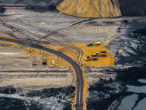 Aerial view of the Garzweiler surface mine with heavy dump trucks and machinery operating on yellow soil and dark coal deposits near a winding road in Juchen, North Rhine-Westphalia, Germany.