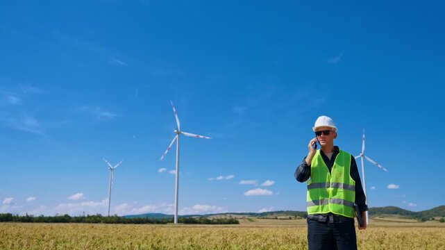 Technician talking on mobile phone at wind farm. A man wearing a reflective vest stands in an open field near wind turbines on a clear day.
