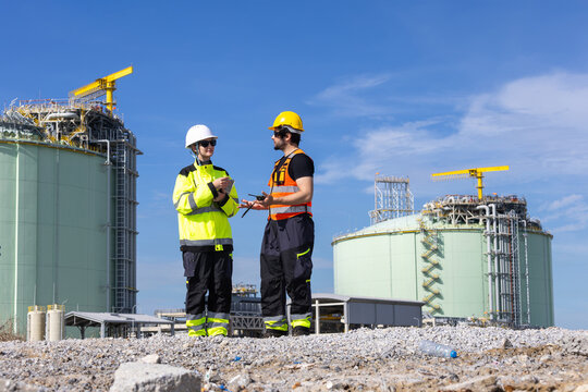 LNG plant engineers discussing project in front of large storage tanks, liquefied natural gas workers in safety gear talking at industrial facility, technician inspecting lng terminal