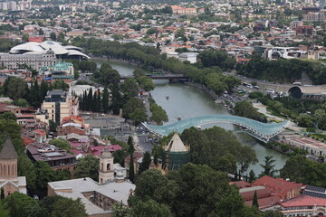 Aerial view of Tbilisi, Georgia with Peace Bridge over Kura river, historic old town, church towers and modern concert hall among trees © Ольга Дербасова