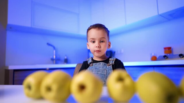 Child sitting behind row of apples at illuminated counter. A boy wears denim overalls while watching yellow fruits arranged on a white table in a blue lit room.
