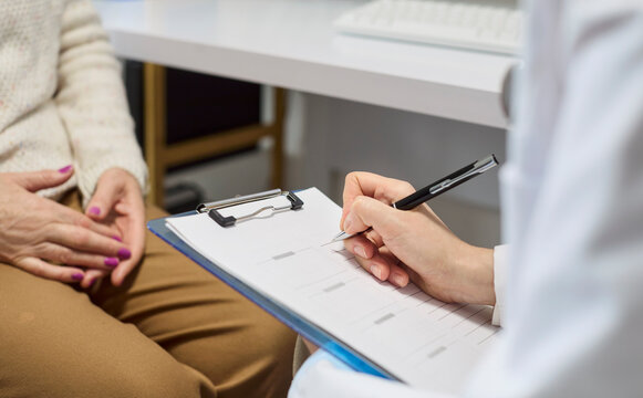 Doctor and senior patient consultation in clinic. Closeup of doctor writing notes on clipboard while listening and preparing diagnosis. Symbolizes trustful medical care and communication.
