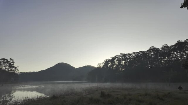 Aerial drone view of a fisherman rowing across Tuyen Lam Lake at sunrise near Suoi Tia in Xuan Huong, Da Lat, Vietnam, surrounded by peaceful pine forests.