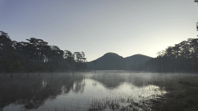 Aerial drone view of a fisherman rowing across Tuyen Lam Lake at sunrise near Suoi Tia in Xuan Huong, Da Lat, Vietnam, surrounded by peaceful pine forests.