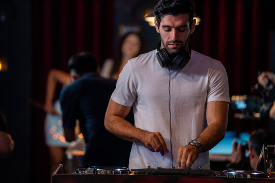 A wide view of a male DJ standing behind turntables and a mixer in a busy nightclub reaches for the pitch fader in a wash of red light in a wide nightclub interior with pool tables.