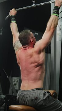 Man performs seated lat pulldown on cable machine in dark gym. Athlete pulls bar toward chest engaging back muscles with steady control