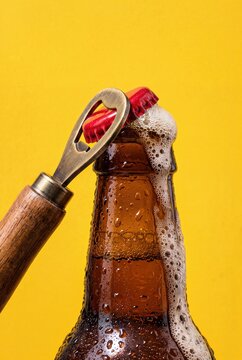 Cold beer bottle opening with metal opener and overflowing foam on yellow background