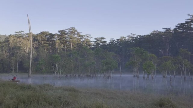 Aerial drone view of a fisherman rowing across Tuyen Lam Lake at sunrise near Suoi Tia in Xuan Huong, Da Lat, Vietnam, surrounded by peaceful pine forests.