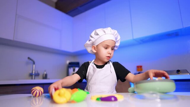 Child chef with toy cookware in blue kitchen. The young cook reaches across colorful pieces arranged on a glossy white table.