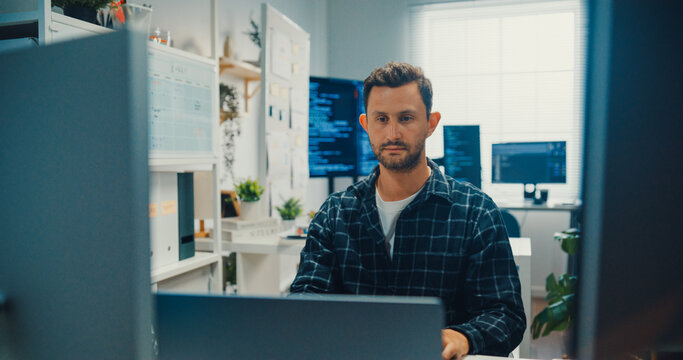 Young Caucasian male programmer writes code using multiple screens in home office workspace. AI development, cybersecurity, network database system, software engineering, remote tech startup concept.