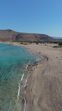 Waves gently lap the shore at Simos Beach, Elafonisos Island, Greece, showcasing stunning turquoise waters and soft sands. Holidaymakers relax under the warm sun