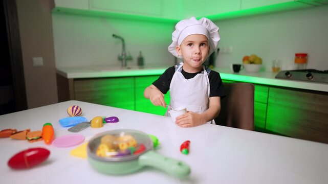 Child chef stirring cup beside toy food. Colorful plastic cookware fills the foreground while green light runs beneath white cabinets.