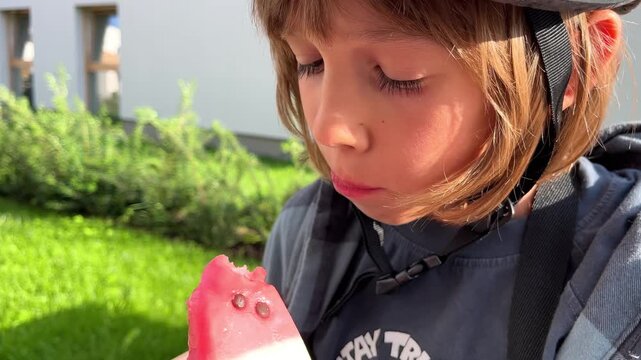 A young boy holding and licking a colorful watermelon-shaped ice pop outdoors on a sunny day, food industry, social media marketing, summer refreshment, sweet treat moment, childhood joy.