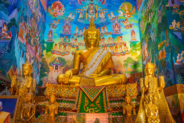 Obraz premium Golden Buddha statue inside the hall of Wat Sri Uthumphon in Mueang Nakhon Sawan District, Nakhon Sawan, Thailand, surrounded by vibrant colorful traditional Thai wall mural painting art.