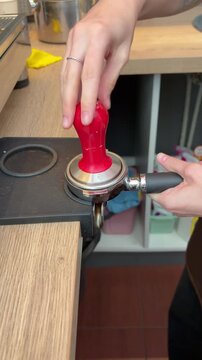A barista tamps ground coffee into a portafilter using a red tamper.