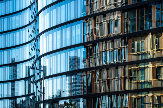 Curved blue glass building facades reflecting neighboring structures and sky, creating layered patterns of modern urban architecture and transparency
