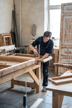 Professional carpenter secures parts of antique wooden window frame to workbench using clamps tools, engaged male crafter carefully fixes instruments to work table, process of restoring old furniture.